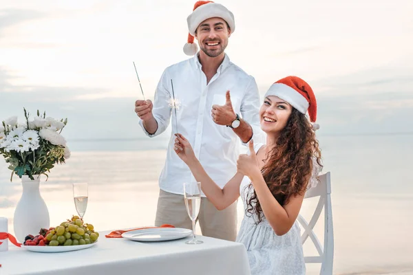 Feliz pareja en sombreros de santa mostrando los pulgares hacia arriba y celebrando la Navidad con bengalas en la playa - foto de stock