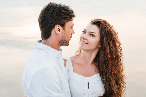 Sonriente pareja romántica mirándose y abrazándose en la orilla del mar - foto de stock