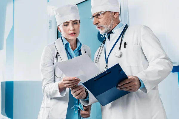 Médicos hombres y mujeres serios mirando el papel y discutiendo en el pasillo del hospital - foto de stock