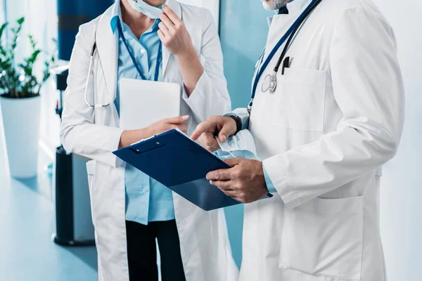 Partial view of male doctor pointing at clipboard to female doctor with digital tablet in hospital corridor — Stock Photo