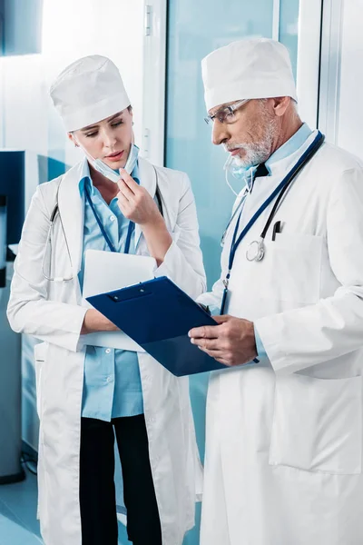 Doctores enfocados en hombres y mujeres mirando el portapapeles y discutiendo en el pasillo del hospital - foto de stock