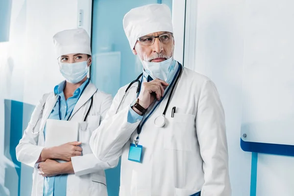 Médico masculino serio quitándose la máscara médica mientras que su colega de pie detrás con la tableta digital en el pasillo del hospital - foto de stock