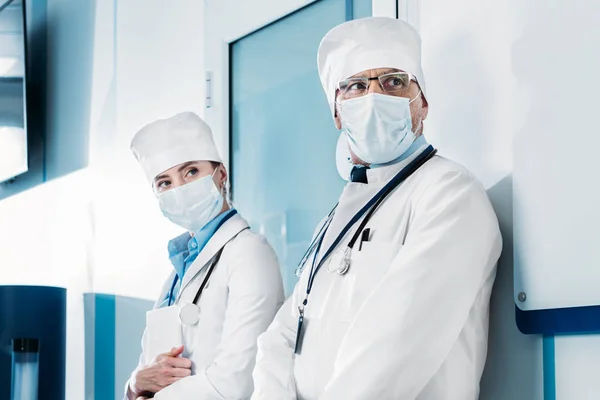Médicos masculinos e femininos em máscaras médicas em pé e olhando para longe no corredor hospitalar — Fotografia de Stock