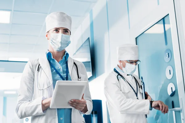 Low angle view of female doctor in medical mask using digital tablet while her male colleague standing behind with clipboard in hospital corridor — Stock Photo