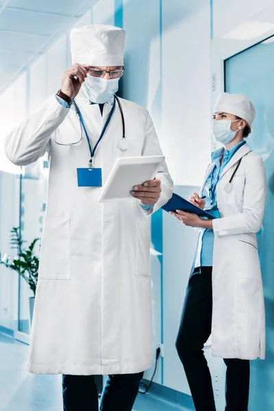 Концентрований лікар-чоловік у медичній масці, що регулює окуляри та використовує цифровий планшет, а його колега-жінка стоїть позаду з кишені в лікарняному коридорі — стокове фото