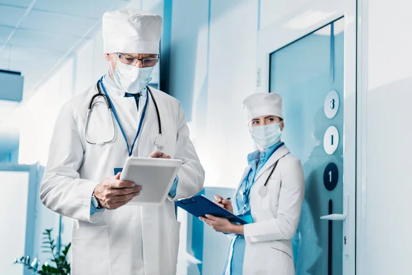 Focused male doctor in medical mask using digital tablet while his female colleague standing behind with clipboard in hospital corridor — Stock Photo