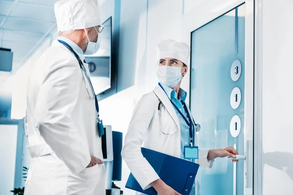 Doctora con portapapeles abriendo la puerta y hablando con un colega en el pasillo del hospital - foto de stock