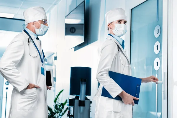 Doctora con portapapeles abriendo la puerta mientras su colega espera en el pasillo del hospital - foto de stock