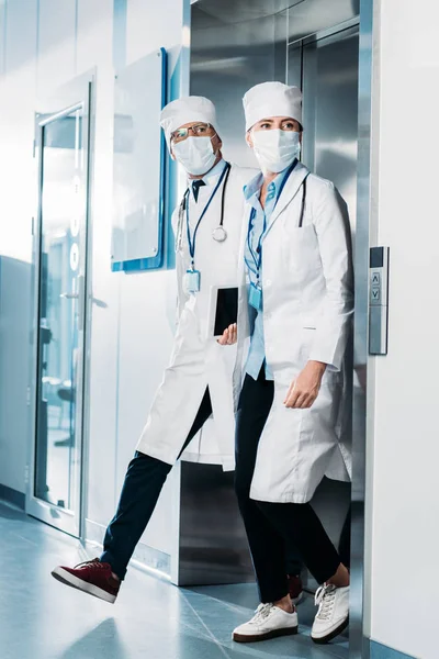 Male and female doctors with digital tablet and clipboard walking out from elevator in hospital corridor — Stock Photo