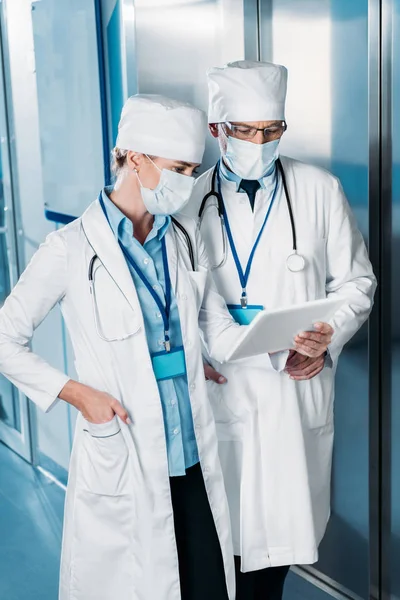 Female doctor in medical mask showing digital tablet to male colleague in hospital corridor — Stock Photo