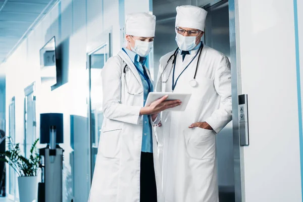 Selective focus of female doctor in medical mask showing digital tablet to male colleague in hospital corridor — Stock Photo