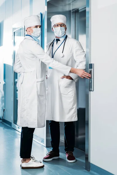 Male and female doctors in medicals masks pushing button of elevator in hospital corridor — Stock Photo