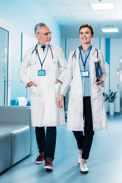 Smiling female and male doctors with badges and stethoscopes over neck walking in hospital corridor — Stock Photo