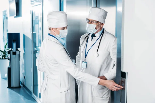 Doctora en máscara médica pulsando el botón del ascensor y hablando con colega masculino en el pasillo del hospital - foto de stock