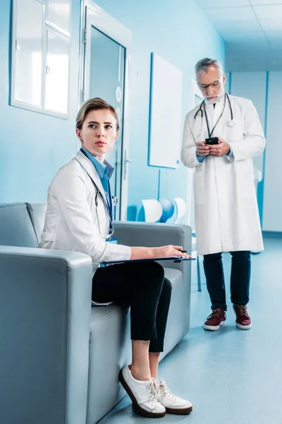Adult female doctor looking away and sitting with clipboard on sofa while her male colleague using smartphone behind in hospital corridor — Stock Photo