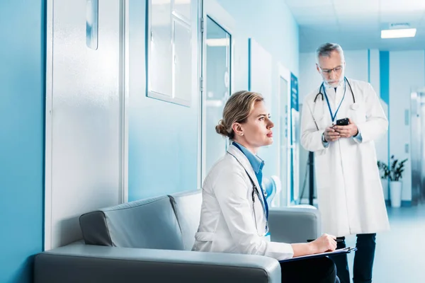 Focused adult female doctor sitting with clipboard on sofa while her male colleague using smartphone behind in hospital corridor — Stock Photo