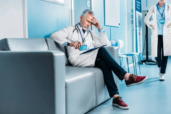 Meditado médico varón de mediana edad con portapapeles sentado en el sofá, mientras que su colega mujer caminando detrás en el pasillo del hospital - foto de stock