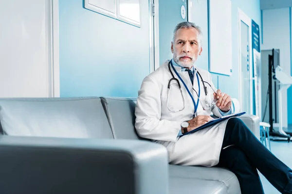 Middle aged male doctor with clipboard looking away and sitting on couch in hospital corridor — Stock Photo