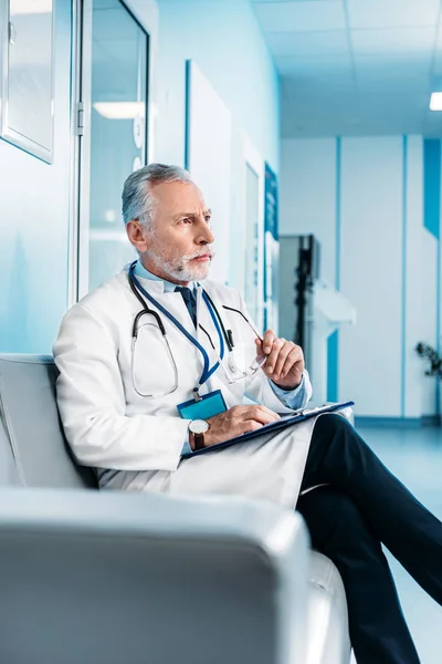 Focused mature male doctor with clipboard holding sitting on couch in hospital corridor — Stock Photo