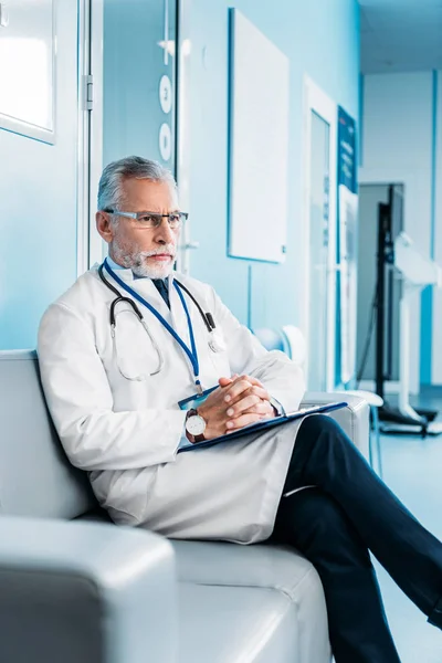 Pensive middle aged male doctor with clipboard sitting on couch in hospital corridor — Stock Photo