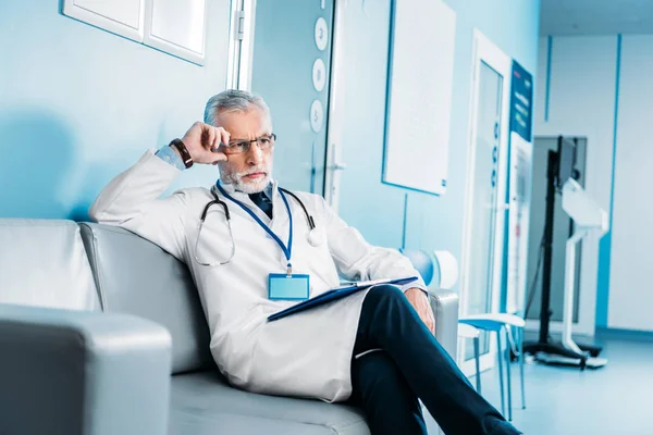 Thoughtful middle aged male doctor with clipboard sitting on couch in hospital corridor — Stock Photo