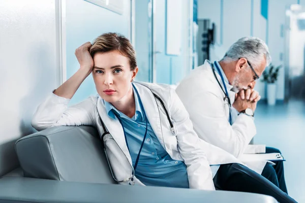 Cansada doctora mirando la cámara en el sofá mientras su colega sentado detrás en el pasillo del hospital - foto de stock