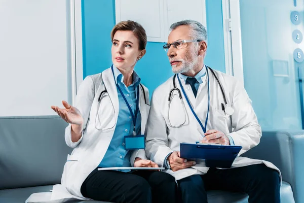 Female doctor with digital tablet pointing by hand to male colleague on sofa in hospital corridor — Stock Photo