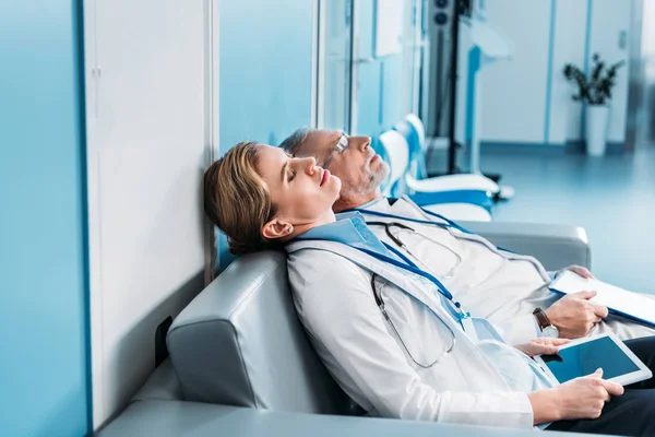 Side view of tired male and female doctors sleeping with clipboard and digital tablet with blank screen on sofa in hospital corridor — Stock Photo