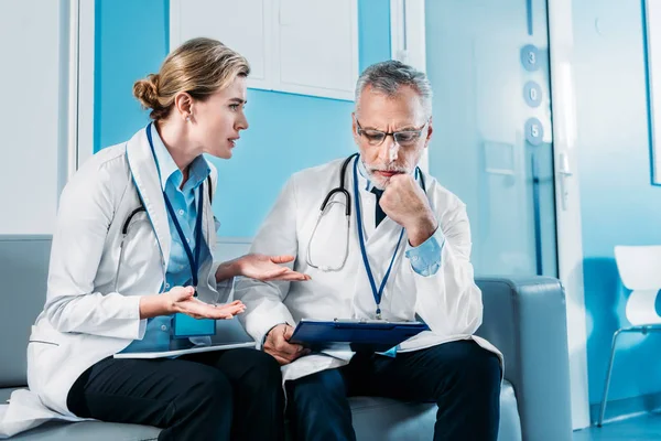 Adult emotional female doctor gesturing by hands and having discussion with male colleague on sofa in hospital corridor — Stock Photo