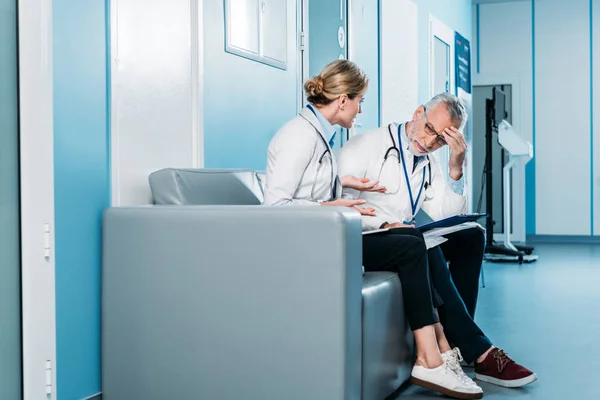 Vista lateral del emotivo médico femenino gesticulando con las manos y discutiendo con un colega masculino en el sofá en el pasillo del hospital - foto de stock