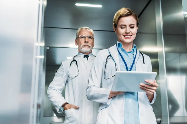 Felice medico femminile utilizzando tablet digitale mentre il suo collega maschio in piedi dietro in ascensore — Foto stock