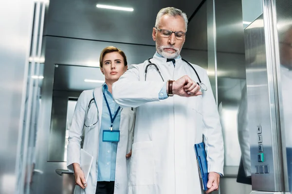 Серйозний чоловік середнього віку лікар перевіряє наручний годинник, а його колега-жінка стоїть позаду в лікарняному ліфті — стокове фото