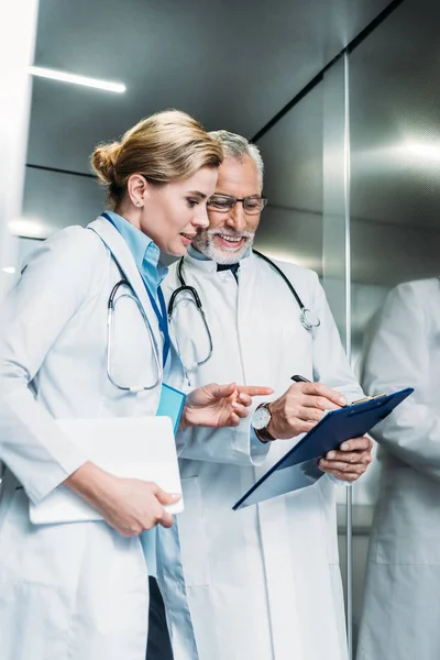 Smiling mature male doctor showing clipboard to female colleague with digital tablet in hospital elevator — Stock Photo