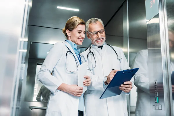 Glücklich reifer männlicher Arzt zeigt Kollegin Klemmbrett mit digitalem Tablet im Krankenhausaufzug — Stockfoto