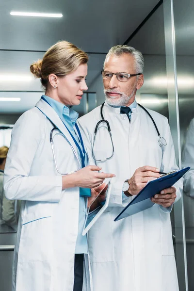 Doctor varón mostrando portapapeles a colega mujer con tableta digital en ascensor del hospital - foto de stock