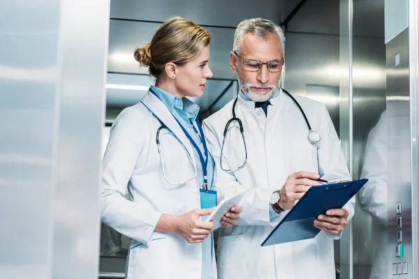 Mature male doctor showing clipboard to female colleague with digital tablet in hospital elevator — Stock Photo