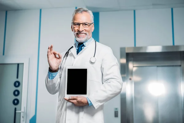 Усміхнений зрілий чоловік лікар зі стетоскопом над шиєю робить нормальний жест і показує цифровий планшет з порожнім екраном в лікарні — стокове фото