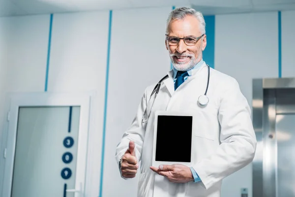 Happy mature male doctor with stethoscope over neck showing thumb up gesture and holding digital tablet with blank screen in hospital — Stock Photo