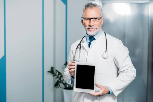 Feliz médico varón maduro con estetoscopio sobre el cuello mostrando tableta digital con pantalla en blanco en el hospital - foto de stock