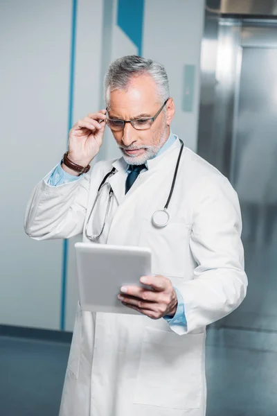 Mature male doctor with stethoscope over neck adjusting eyeglasses and looking at digital tablet in hospital — Stock Photo