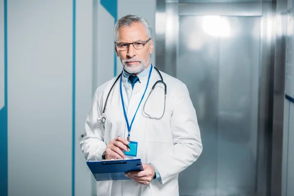 Confident mature male doctor with stethoscope over neck holding clipboard and looking at camera in hospital — Stock Photo