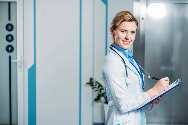Doctora sonriente con estetoscopio sobre el cuello escribiendo en portapapeles en el hospital - foto de stock