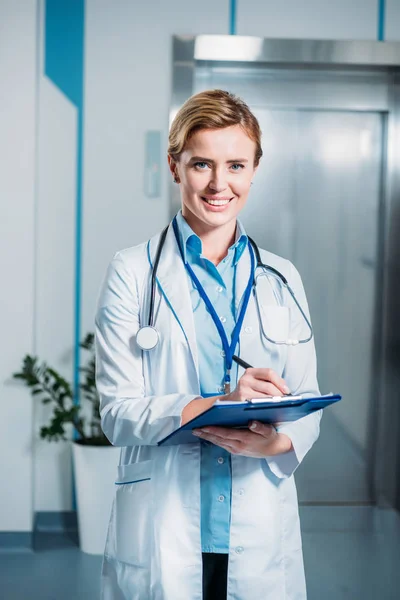 Feliz médico femenino con estetoscopio sobre la escritura del cuello en portapapeles y mirando la cámara en el hospital - foto de stock