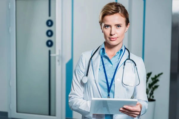 Bellissimo medico femminile adulto con stetoscopio sul collo utilizzando tablet digitale e guardando la fotocamera in ospedale — Stock Photo