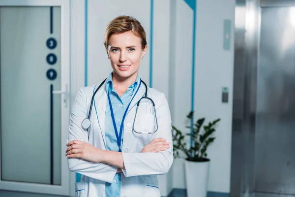 Atractiva doctora sonriente con brazos cruzados mirando a la cámara en el hospital - foto de stock