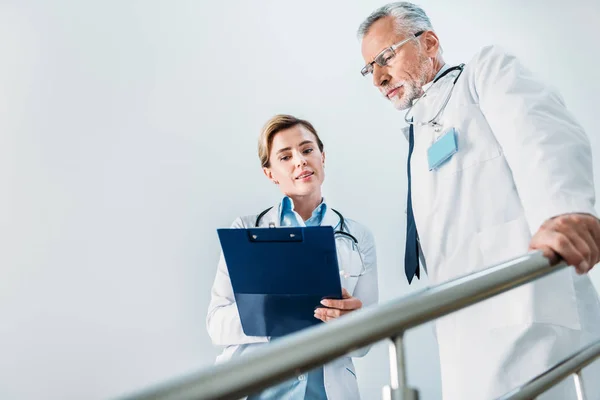 Attractive female doctor showing clipboard to serious male colleague on staircase in hospital — Stock Photo