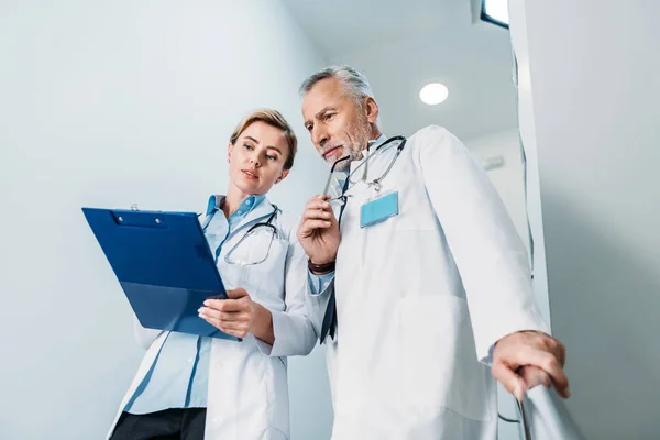 Vista de ángulo bajo del médico femenino que muestra el portapapeles al colega masculino en la escalera en el hospital - foto de stock