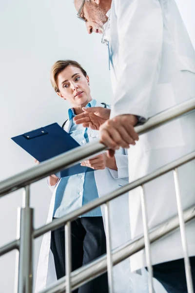 Низький кут зору жінки-лікаря, що розмовляє і показує буфер обміну чоловічому колезі на сходах у лікарні — стокове фото