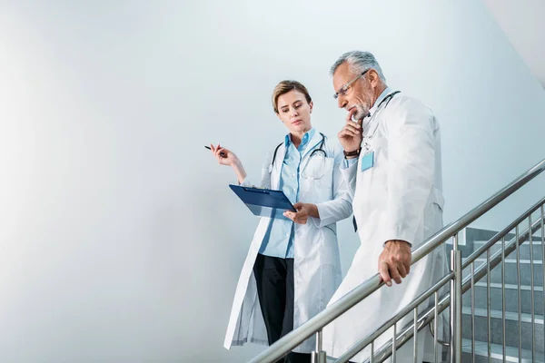 Adult female doctor showing clipboard to thoughtful male colleague on staircase in hospital — Stock Photo