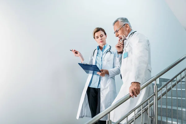 Doctora adulta mostrando portapapeles a un colega varón reflexivo en la escalera en el hospital - foto de stock